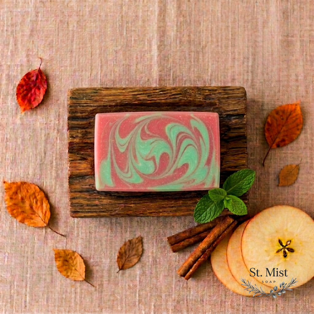 St. Mist Soap Apple Cider bar with red and green swirls on a wooden dish, surrounded by autumn leaves and apple slices.
