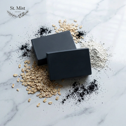 St. Mist Soap black charcoal and oatmeal artisan bar soap on a marble surface with raw ingredients.