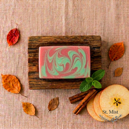 St. Mist Soap Apple Cider bar with red and green swirls on a wooden dish, surrounded by autumn leaves and apple slices.