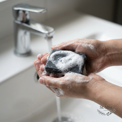 St. Mist Soap handcrafted black charcoal and oatmeal soap bar creating a rich, creamy lather in hands under a faucet.